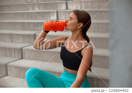Fit woman enjoying a refreshing drink while resting on outdoor steps, showcasing healthy lifestyle and fitness dedication in vibrant attire. Workout concept Fit woman enjoying a refreshing drink while resting on outdoor steps, showcasing healthy lifestyle and fitness dedication in vibrant attire. Workout concept 128804402