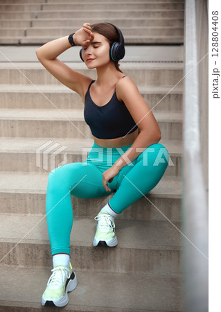 Stylish fitness model relaxes on stairs, sipping water from a bottle, embodying a healthy outdoor lifestyle and dedication to fitness. Workout concept. Stylish fitness model relaxes on stairs, sipping water from a bottle, embodying a healthy outdoor lifestyle and dedication to fitness. Workout concept. 128804408