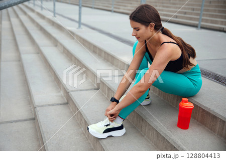 Fit woman tying shoelaces while sitting on outdoor steps, wearing athletic gear, preparing for an energetic workout session in nature. Workout concept 128804413