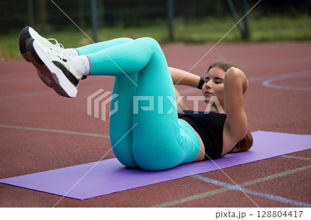 Fit woman performing crunches on a yoga mat outdoors, showcasing strength and determination in a vibrant athletic outfit during exercise. Workout concept 128804417