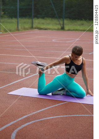 Fit woman stretching on a yoga mat outdoors, showcasing flexibility and strength in a vibrant athletic outfit during exercise routine. Workout concept 128804425
