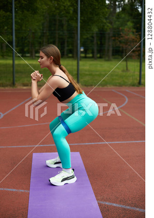 Fit woman performing squats outdoors on a purple mat, showcasing strength and determination in a vibrant green park environment. Workout concept 128804432