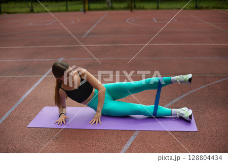 Fit woman exercising outdoors on a purple mat, performing side leg lifts with resistance band, showcasing strength and determination in fitness. Workout concept 128804434