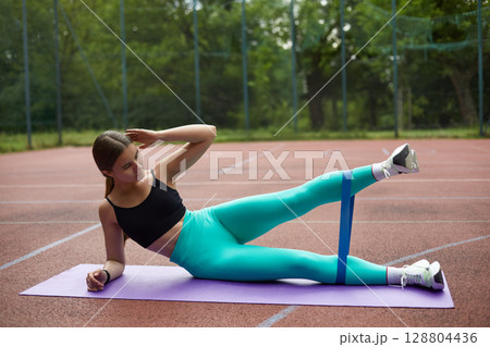 Fit woman exercising outdoors on a yoga mat, performing leg lifts with resistance band, showcasing strength and dedication to fitness. Workout concept Fit woman exercising outdoors on a yoga mat, performing leg lifts with resistance band, showcasing strength and dedication to fitness. Workout concept 128804436