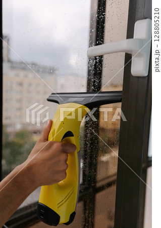 Close-up of male hand using cordless vacuum cleaner for cleaning window, wiping away water drops on rainy day, ensuring spotless and hygienic indoor environment. Concept of household cleanliness. Close-up of male hand using cordless vacuum cleaner for cleaning window, wiping away water drops on rainy day, ensuring spotless and hygienic indoor environment. Concept of household cleanliness. 128805210