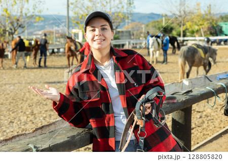 Skilled European young horsewoman in plaid shirt holding harness in hands during work on ranch 128805820