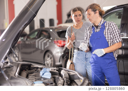 Woman talking to auto mechanic in uniform about repairing his car engine in auto repair shop 128805860