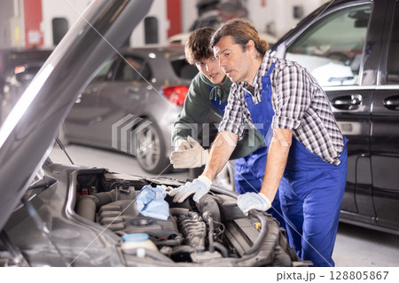 Two mechanics examining car engine under open hood in workshop 128805867