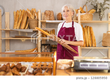 Mature saleswoman holding paper bag with baguettes in bakery Mature saleswoman holding paper bag with baguettes in bakery 128805950