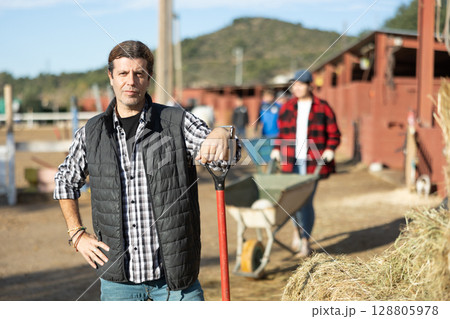 Skilled adult man stables worker in plaid shirt looking at camera while holding working tool on horse ranch in spring Skilled adult man stables worker in plaid shirt looking at camera while holding working tool on horse ranch in spring 128805978
