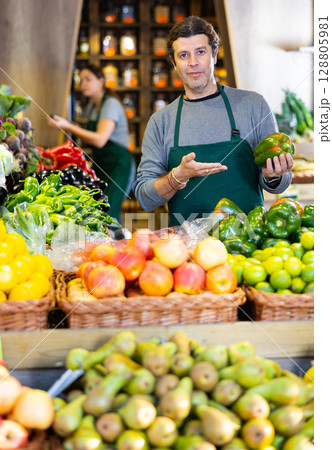 Male seller holding green bell peppers standing in fruit and vegetable section of supermarket 128805981