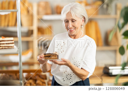 Pleased mature woman holding pastry on paper plate in bakery 128806049