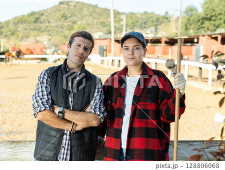 Experienced man and woman in plaid shirts looking at camera while holding working tool on horse ranch in spring 128806068