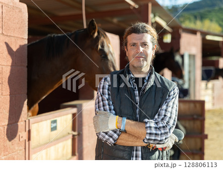 Portrait of owner of horse farm against the background of stall with horses Portrait of owner of horse farm against the background of stall with horses 128806113