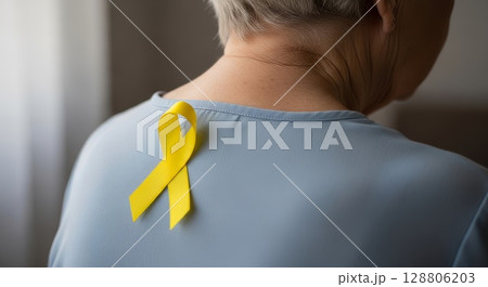 Close-up back view of European woman wearing light blue shirt with bright yellow awareness ribbon pinned on shoulder blade area 128806203