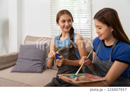 Lesbian couple sharing laughter while painting together indoors 128807957