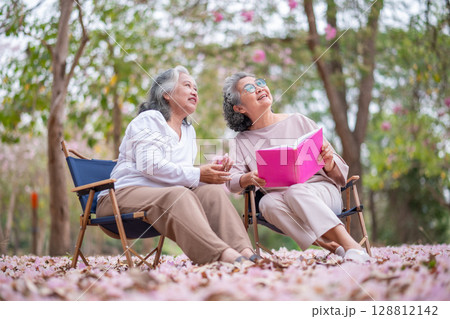 Elderly women under tree adorned pink blossoms, reading book heartwarming and inspiring representation of senior women embracing technology, nature lifestyle. 128812142