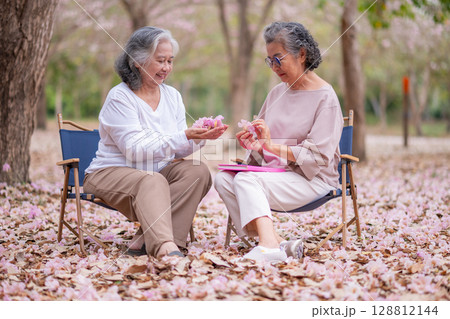 Elderly women under tree adorned pink blossoms, reading book heartwarming and inspiring representation of senior women embracing technology, nature lifestyle. 128812144