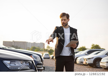 A car salesman in a suit points to a vehicle while holding a tablet in a parking lot on a sunny day, demonstrating a car sale. A car salesman in a suit points to a vehicle while holding a tablet in a parking lot on a sunny day, demonstrating a car sale. 128812192