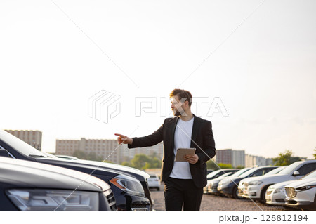 A businessman in a suit points towards a car in a parking lot, holding a tablet while reviewing the vehicles on display. A businessman in a suit points towards a car in a parking lot, holding a tablet while reviewing the vehicles on display. 128812194