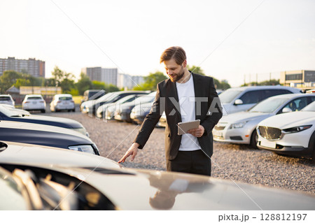 A car salesman checks a car at a dealership. He is holding a tablet while looking at the car with a smile, indicating a positive experience. 128812197