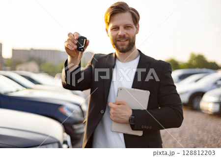 A businessman holds up a car key, smiling at the camera. He appears to be at a car dealership, with cars in the background. 128812203