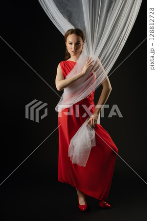 Woman in a flowing red dress poses with sheer fabric in a studio setting, expressing emotion and elegance during a photoshoot Woman in a flowing red dress poses with sheer fabric in a studio setting, expressing emotion and elegance during a photoshoot 128812728