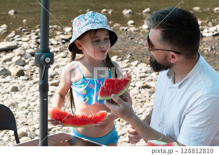 Father and daughter enjoy a refreshing watermelon snack by the river. Father and daughter enjoy a refreshing watermelon snack by the river. 128812810