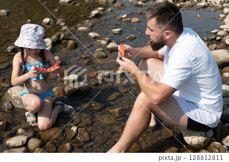A father and daughter enjoy a refreshing watermelon snack by a shallow river on a sunny day. 128812811