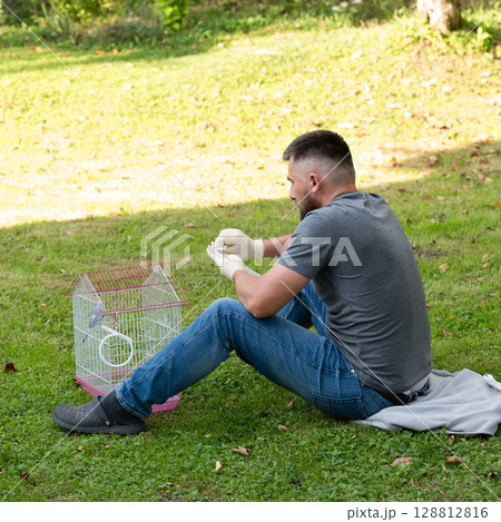 Man gently handles bird outside. Peaceful park setting, bird in cage nearby. 128812816