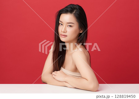 A beautiful young Asian woman with clean, fresh skin against a red background. Skincare, facial treatment, cosmetology, beauty, and spa. An elegant portrait of an Asian woman. 128819450