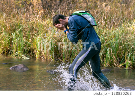 male runner wading through river refreshes his face with water during marathon 128820268