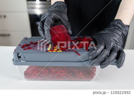 Hands in black gloves expertly grate a vibrant red beetroot using a kitchen shredder for efficient and healthy fresh meal preparation 128822892