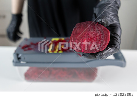 Person in black gloves holds a vibrant, freshly cut beetroot half above a vegetable slicer, illustrating concepts of healthy home cooking Person in black gloves holds a vibrant, freshly cut beetroot half above a vegetable slicer, illustrating concepts of healthy home cooking 128822893