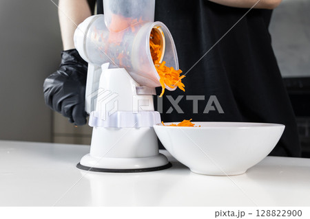 Home chef utilizing a manual kitchen grater for efficiently shredding fresh orange carrots into a white bowl, ideal for healthy meal preparation 128822900