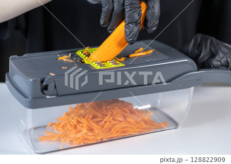 Person's gloved hand expertly grates fresh orange carrot on a modern kitchen slicer, showcasing efficient meal preparation and healthy cooking 128822909