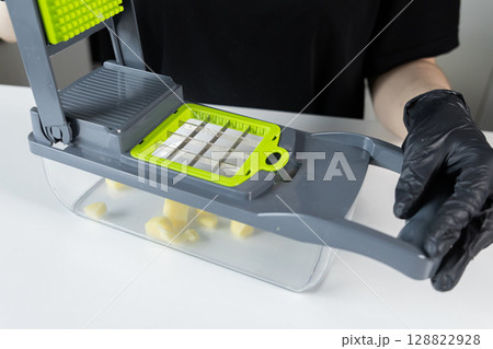 Gloved person uses a modern grey and green kitchen vegetable chopper, efficiently dicing raw potatoes for quick food preparation and convenient healthy cooking 128822928
