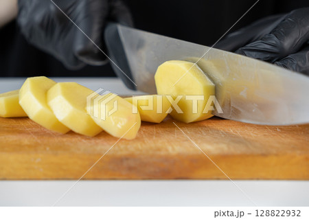 Close-up of a chef's hands in black gloves precisely slicing fresh peeled potatoes on a rustic wooden cutting board, ideal for home cooking or professional culinary preparation Close-up of a chef's hands in black gloves precisely slicing fresh peeled potatoes on a rustic wooden cutting board, ideal for home cooking or professional culinary preparation 128822932
