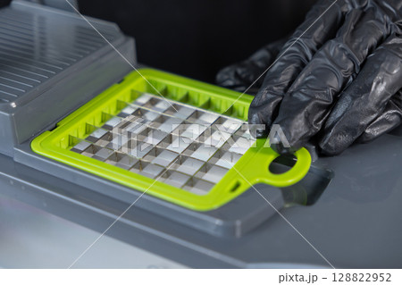 A gloved hand adjusts the vibrant green and metal grid blade insert of a kitchen vegetable chopper, promoting efficient food preparation A gloved hand adjusts the vibrant green and metal grid blade insert of a kitchen vegetable chopper, promoting efficient food preparation 128822952
