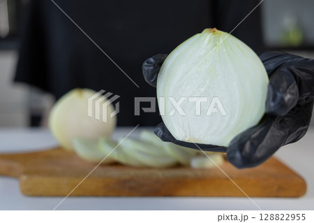 A professional chef's gloved hand presents a freshly sliced white onion half, showcasing crisp layers for healthy culinary dishes on a rustic wooden kitchen cutting board A professional chef's gloved hand presents a freshly sliced white onion half, showcasing crisp layers for healthy culinary dishes on a rustic wooden kitchen cutting board 128822955