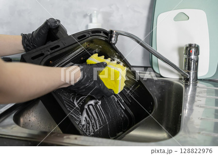 A person wearing protective black gloves diligently cleans a greasy oven baking tray with a soapy yellow sponge, promoting excellent kitchen hygiene and household cleanliness 128822976