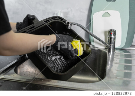 A person wearing protective black gloves diligently cleans an air fryer basket in a kitchen sink, promoting home hygiene and modern appliance care 128822978