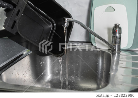 Person wearing black glove pouring dirty water from air fryer basket into a kitchen sink, illustrating effective appliance cleaning Person wearing black glove pouring dirty water from air fryer basket into a kitchen sink, illustrating effective appliance cleaning 128822980