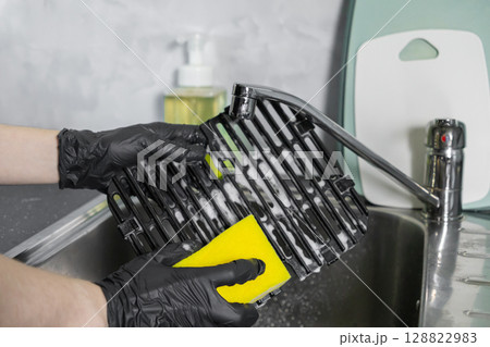 Person in black gloves washing a dirty grill grate with a yellow sponge and soapy water in a kitchen sink, emphasizing home hygiene and cleaning 128822983