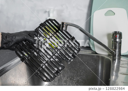 Person wearing a black glove diligently washing a dirty air fryer grill grate covered in foamy soap suds within a clean stainless steel kitchen sink, showcasing essential domestic hygiene 128822984
