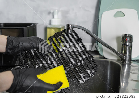 Hands in black gloves diligently cleaning an air fryer grill grate with a yellow sponge and soap in a kitchen sink, for household hygiene 128822985