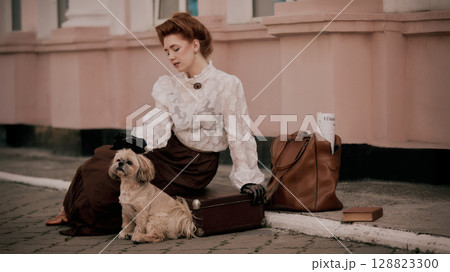 Woman in vintage outfit sitting with dog beside suitcase and leather bag Woman in vintage outfit sitting with dog beside suitcase and leather bag 128823300