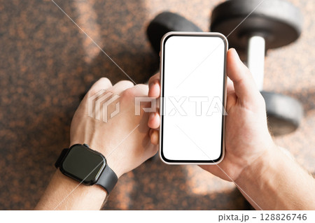 Mans hand holding smartphone with blank screen and wearing smartwatch on wrist, set against background at fitness center. Check fitness progress. Mock up. 128826746