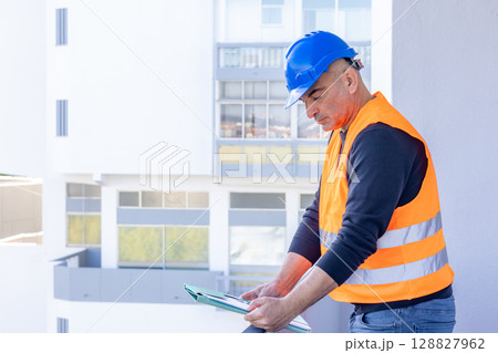 Absorbed construction engineer or building contractor wearing a blue protective helmet checking office blueprints. Copy space 128827962