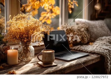 Cozy autumn workspace with laptop, coffee, and candle on wooden desk by window, surrounded by fall foliage Cozy autumn workspace with laptop, coffee, and candle on wooden desk by window, surrounded by fall foliage 128827972